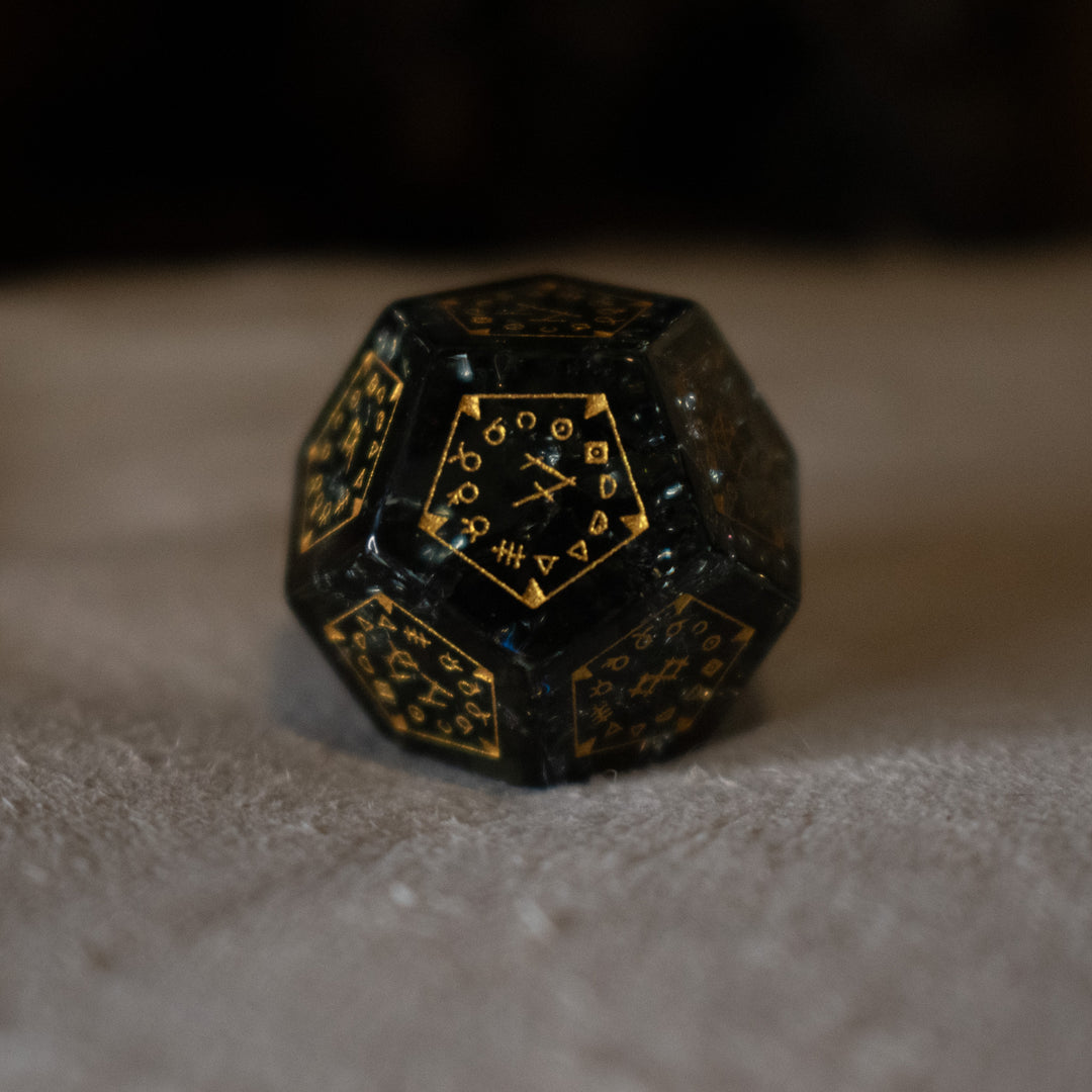 hand-carved, smoky-gray glass dice with gold engraved numbers and wendigo inspired details, displayed against a black background.