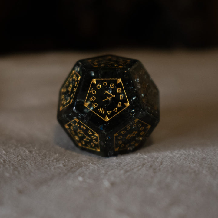 hand-carved, smoky-gray glass dice with gold engraved numbers and wendigo inspired details, displayed against a black background.