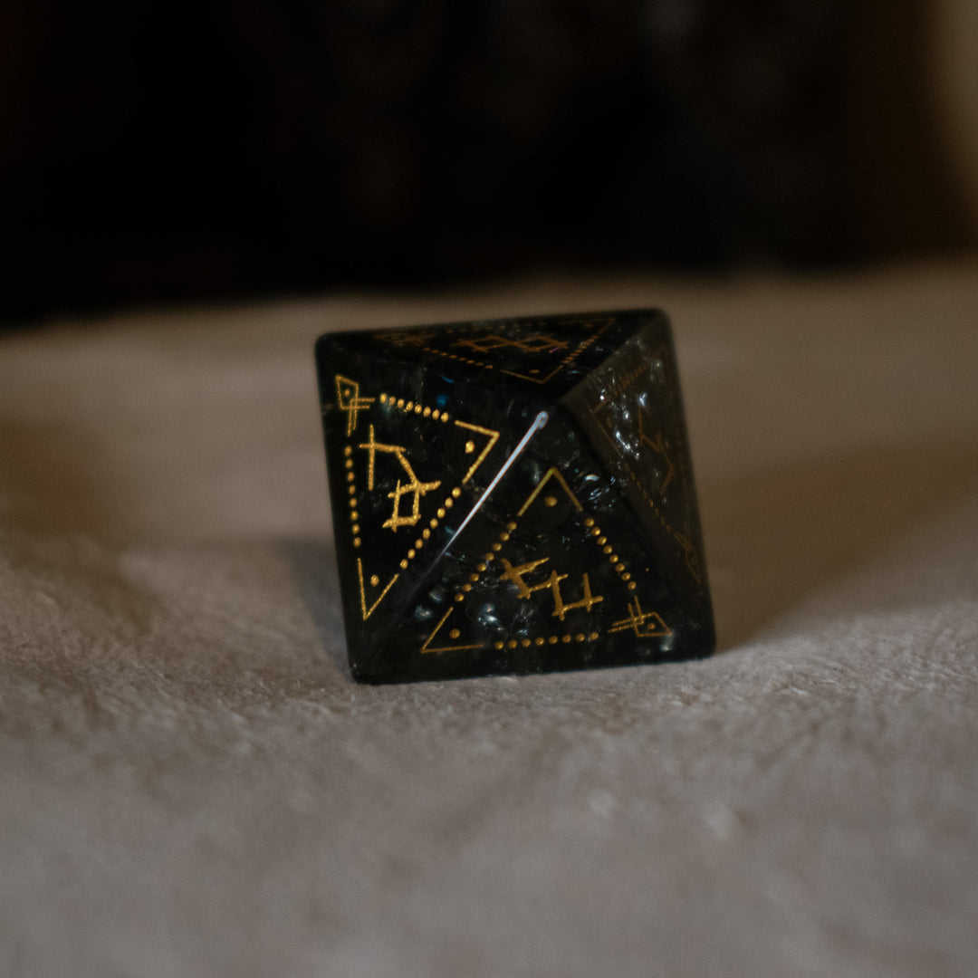 hand-carved, smoky-gray glass dice with gold engraved numbers and wendigo inspired details, displayed against a black background.