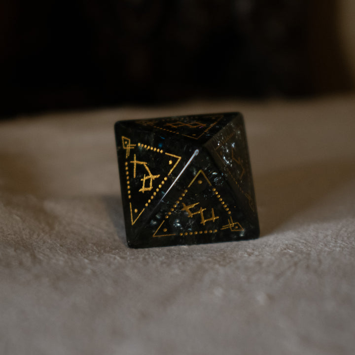 hand-carved, smoky-gray glass dice with gold engraved numbers and wendigo inspired details, displayed against a black background.
