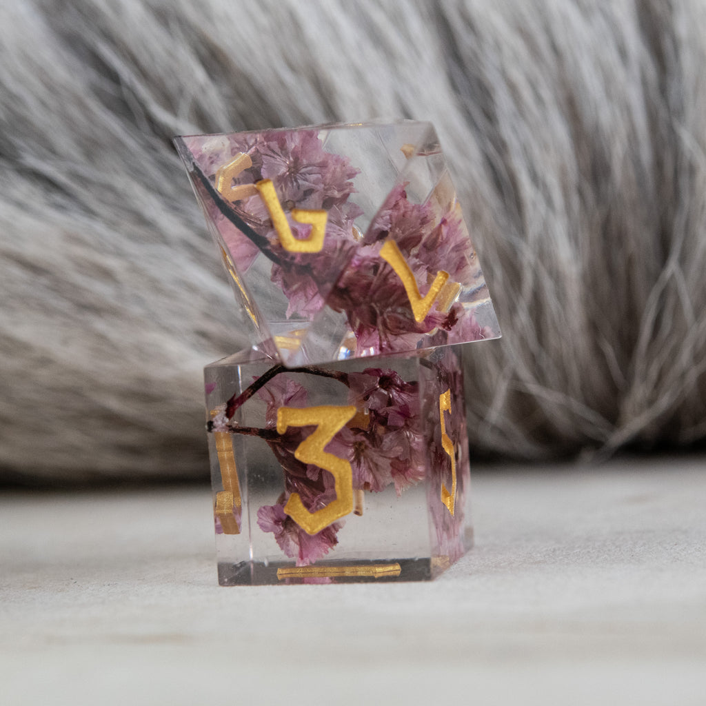 A set of clear sharp-edged resin dice with hand-painted gold numbers, featuring pink florals inside for dungeons and dragons and tabletop gaming