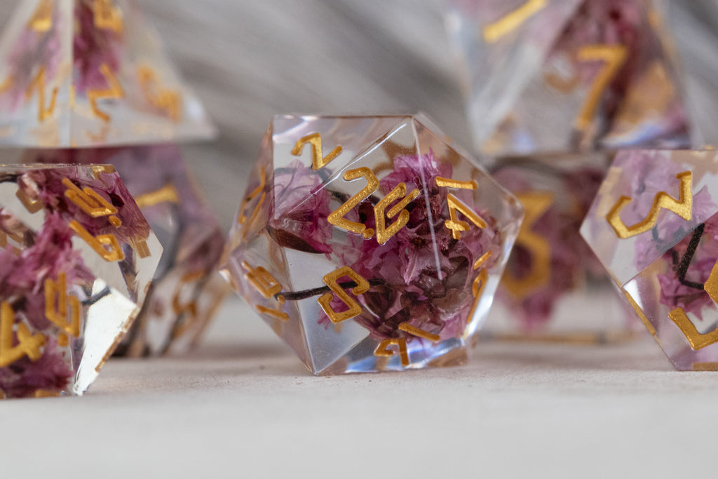 A set of clear sharp-edged resin dice with hand-painted gold numbers, featuring pink florals inside for dungeons and dragons and tabletop gaming
