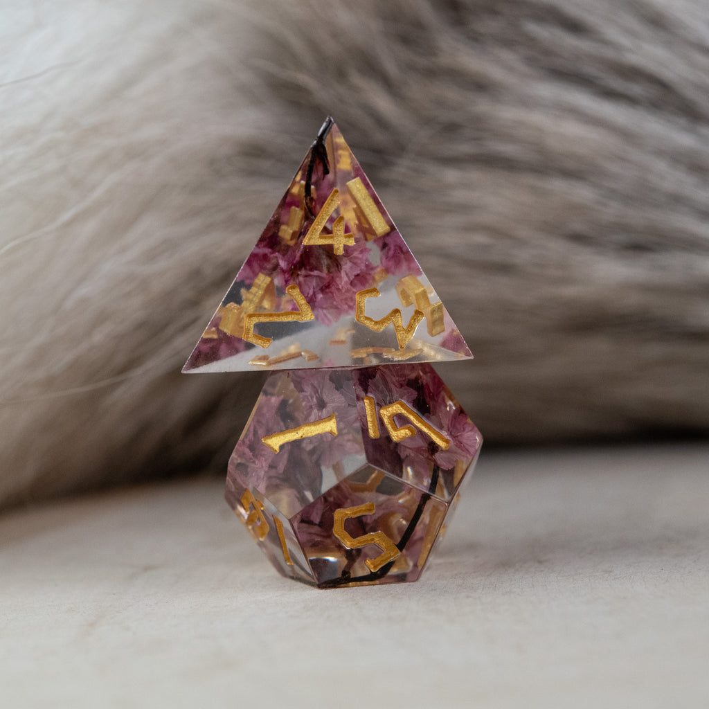 A set of clear sharp-edged resin dice with hand-painted gold numbers, featuring pink florals inside for dungeons and dragons and tabletop gaming