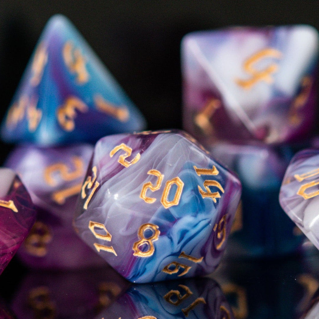 A set of acrylic dice with engraved gold numbers, featuring a mix of purple, blue, and white colors, displayed on a reflective surface.