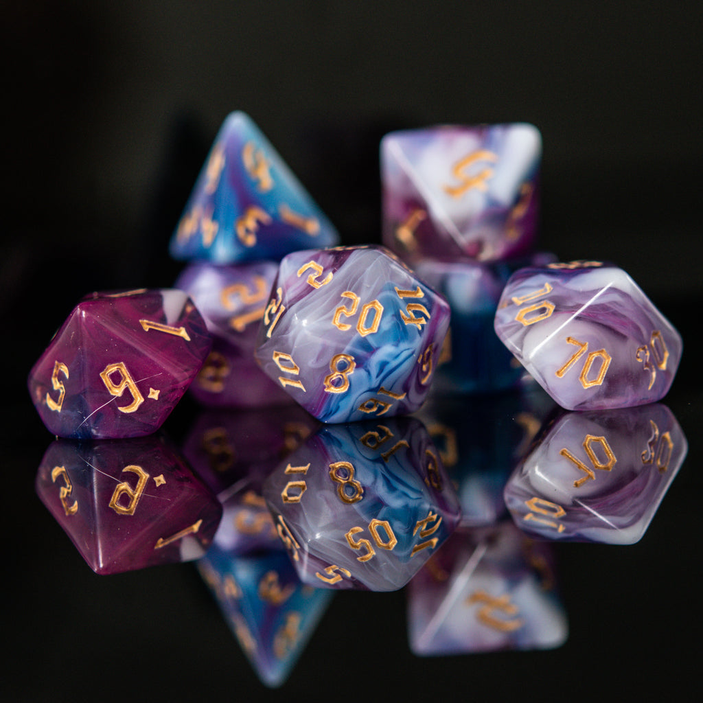 A set of acrylic dice with engraved gold numbers, featuring a mix of purple, blue, and white colors, displayed on a reflective surface.