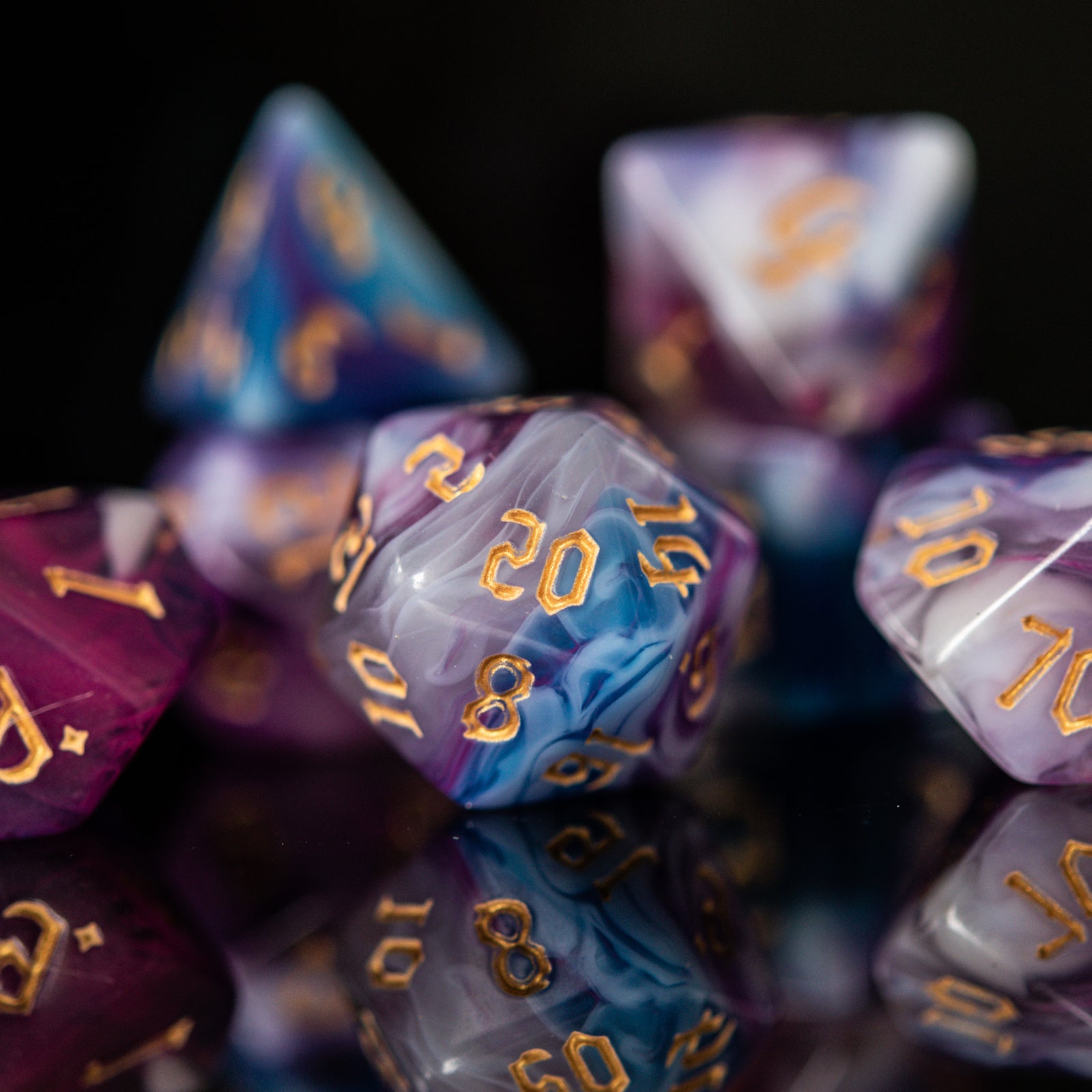 A set of acrylic dice with engraved gold numbers, featuring a mix of purple, blue, and white colors, displayed on a reflective surface.