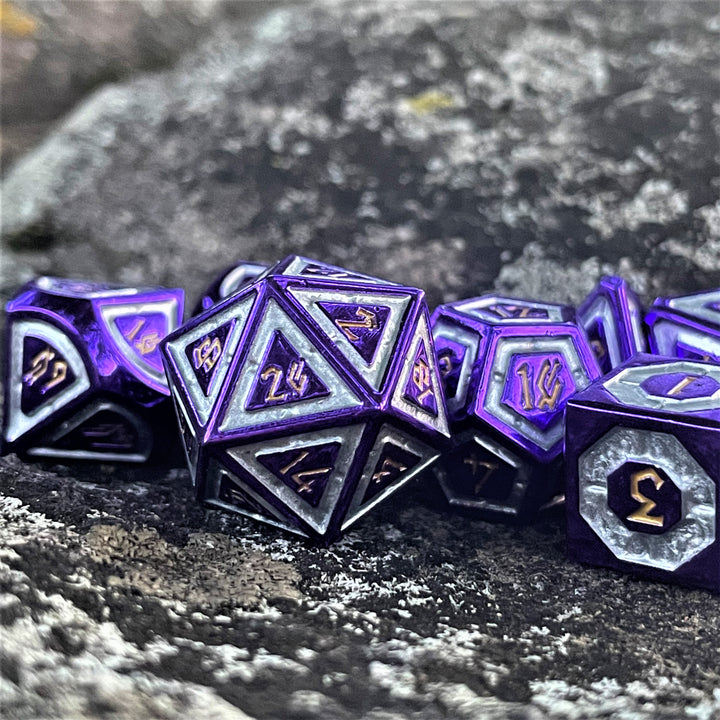 A set of purple and silver metal dice with engraved numbers, including various shapes such as a d4, d6, d8, d10, d12, and d20, displayed on a rocky surface.