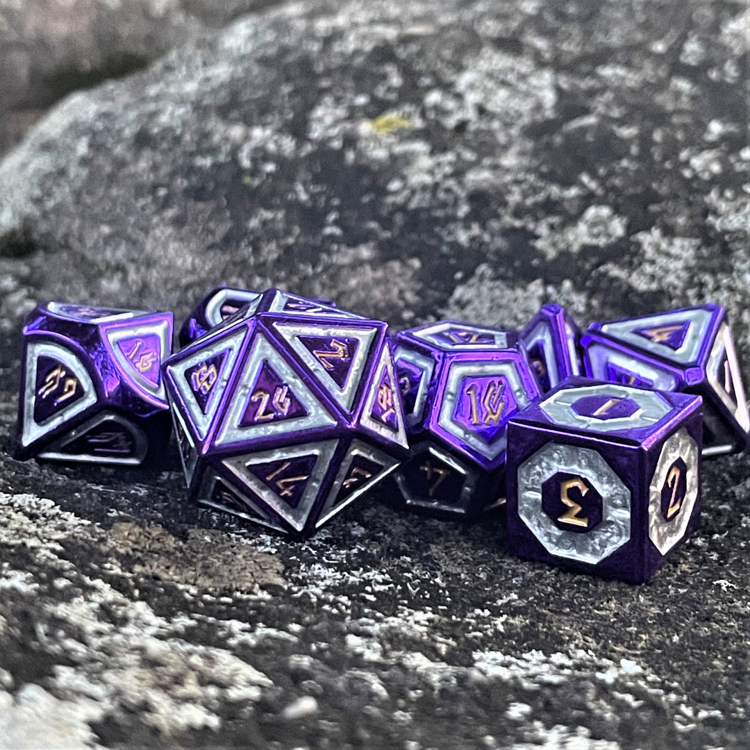 A set of purple and silver metal dice with engraved numbers, including various shapes such as a d4, d6, d8, d10, d12, and d20, displayed on a rocky surface.