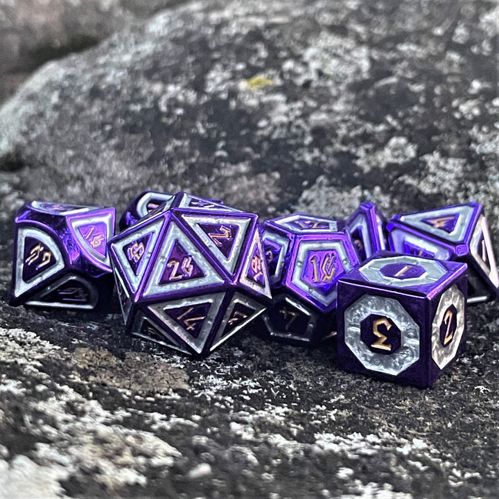 A set of purple and silver metal dice with engraved numbers, including various shapes such as a d4, d6, d8, d10, d12, and d20, displayed on a rocky surface.