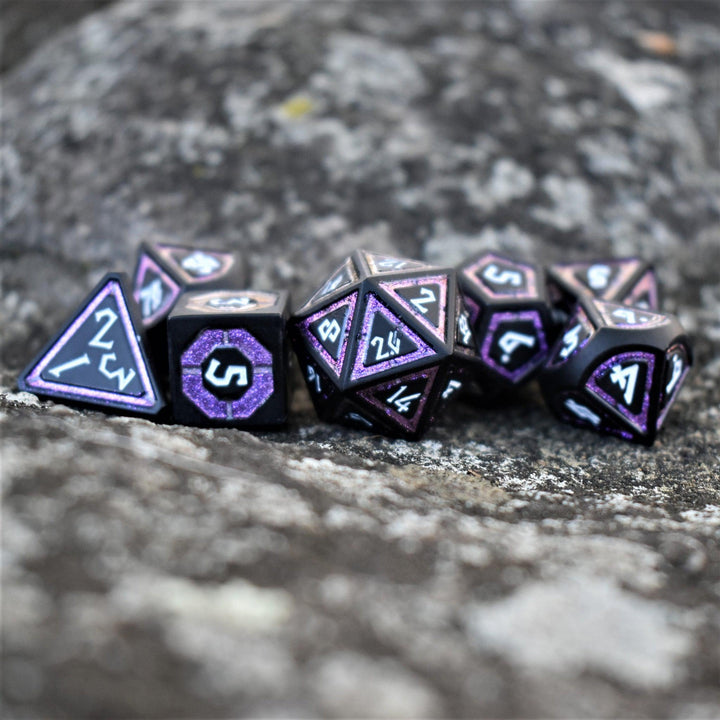 A set of purple and matte black metal dice with engraved numbers on a textured stone surface.