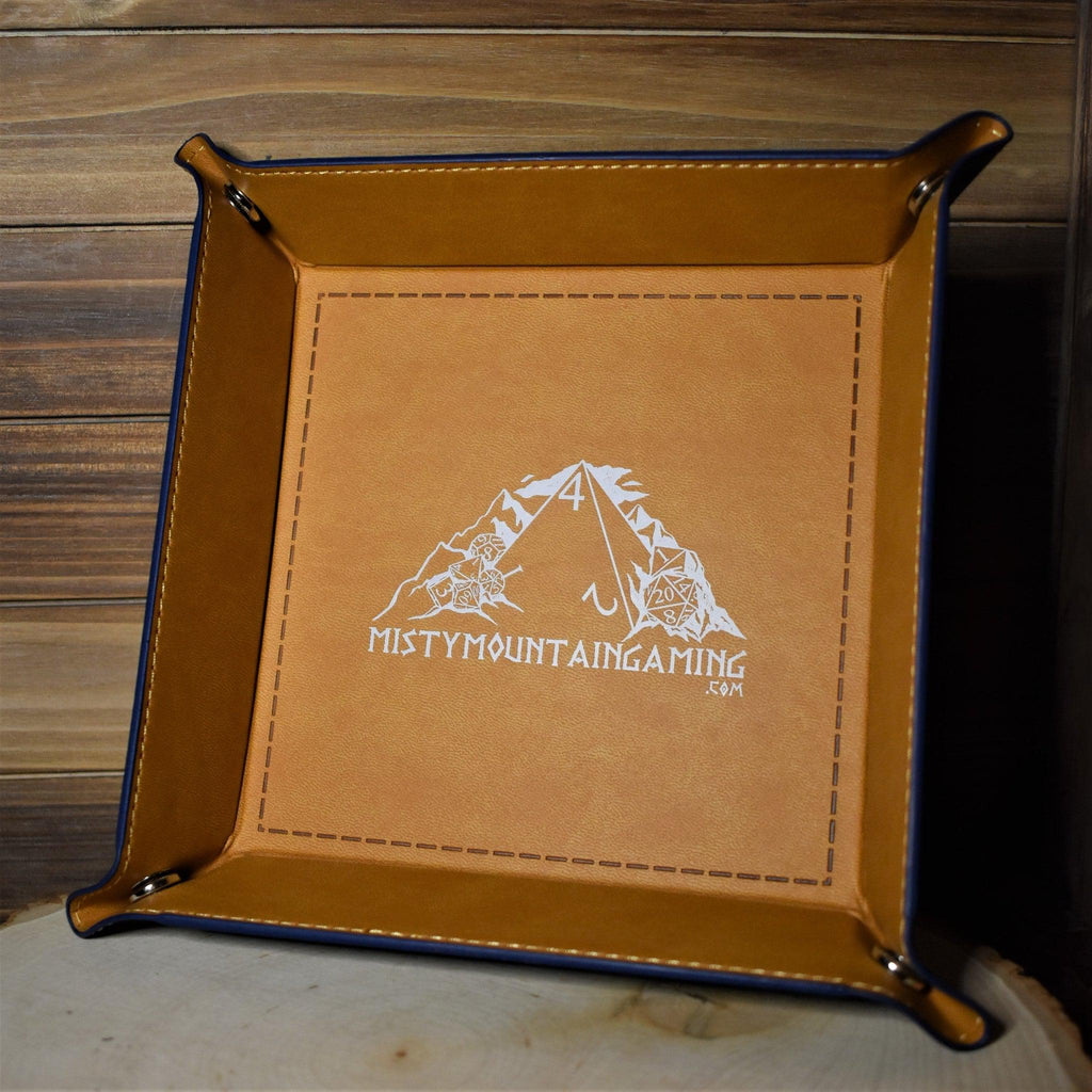 A collapsible dice tray made of poly-leather, featuring the Misty Mountain Gaming logo, placed on a wooden surface.