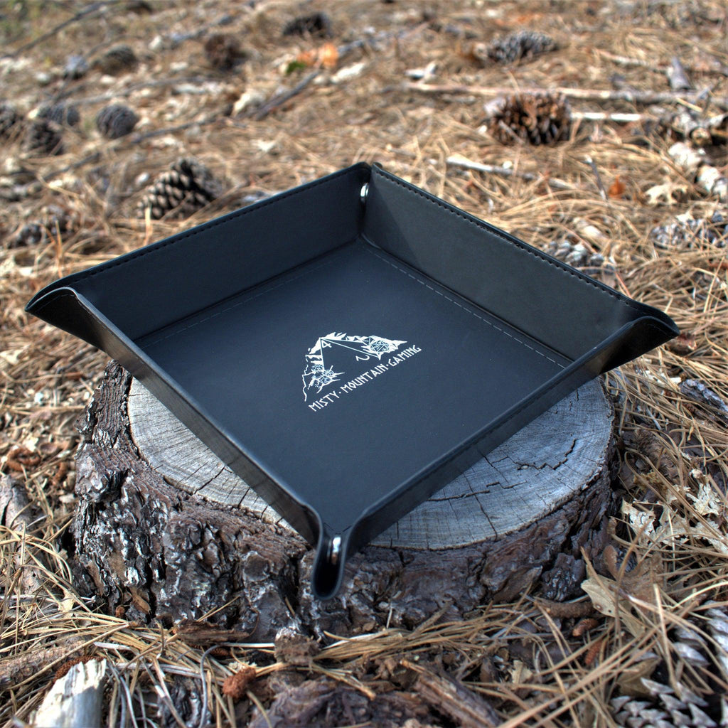 A collapsible dice tray made of poly-leather, featuring the Misty Mountain Gaming logo, placed on a wooden surface.