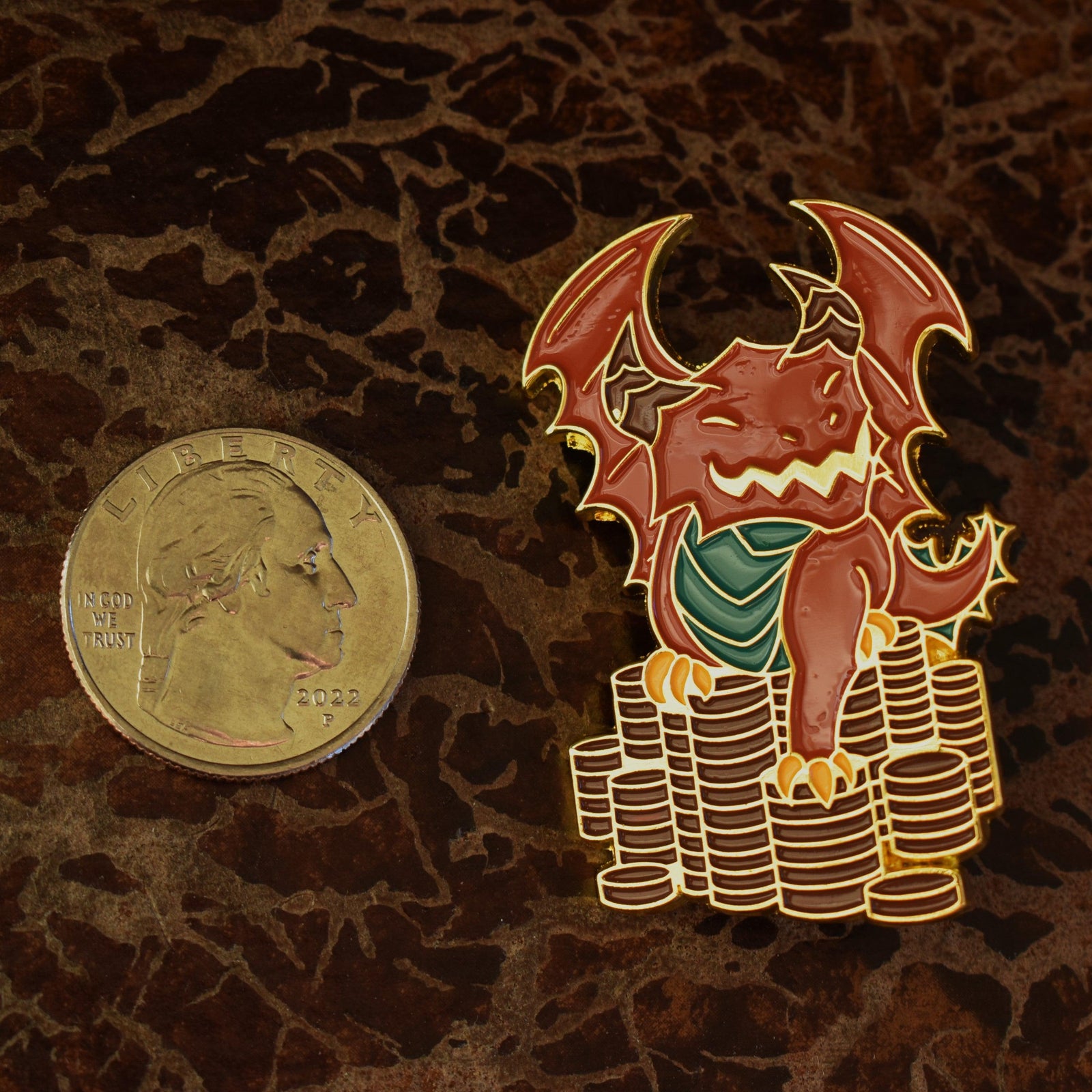 A metal enamel pin shaped like a red dnd dragon sitting on top of a stack of gold coins.