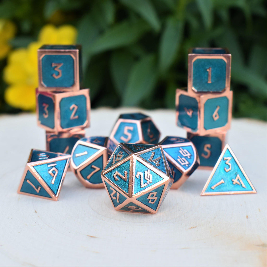 A set of metal dice in bronze and blue colors, including different shapes for a role-playing game. The set is displayed on a white surface with flowers in the background.