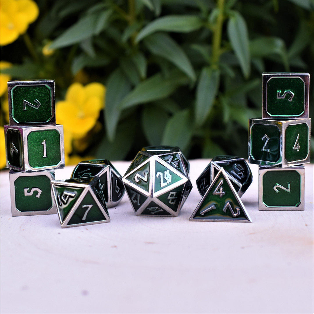 A set of green metal dice with embossed numbers, including various shapes such as a D4, D6, D8, D10, D12, and D20, displayed on a white surface with yellow flowers in the background.