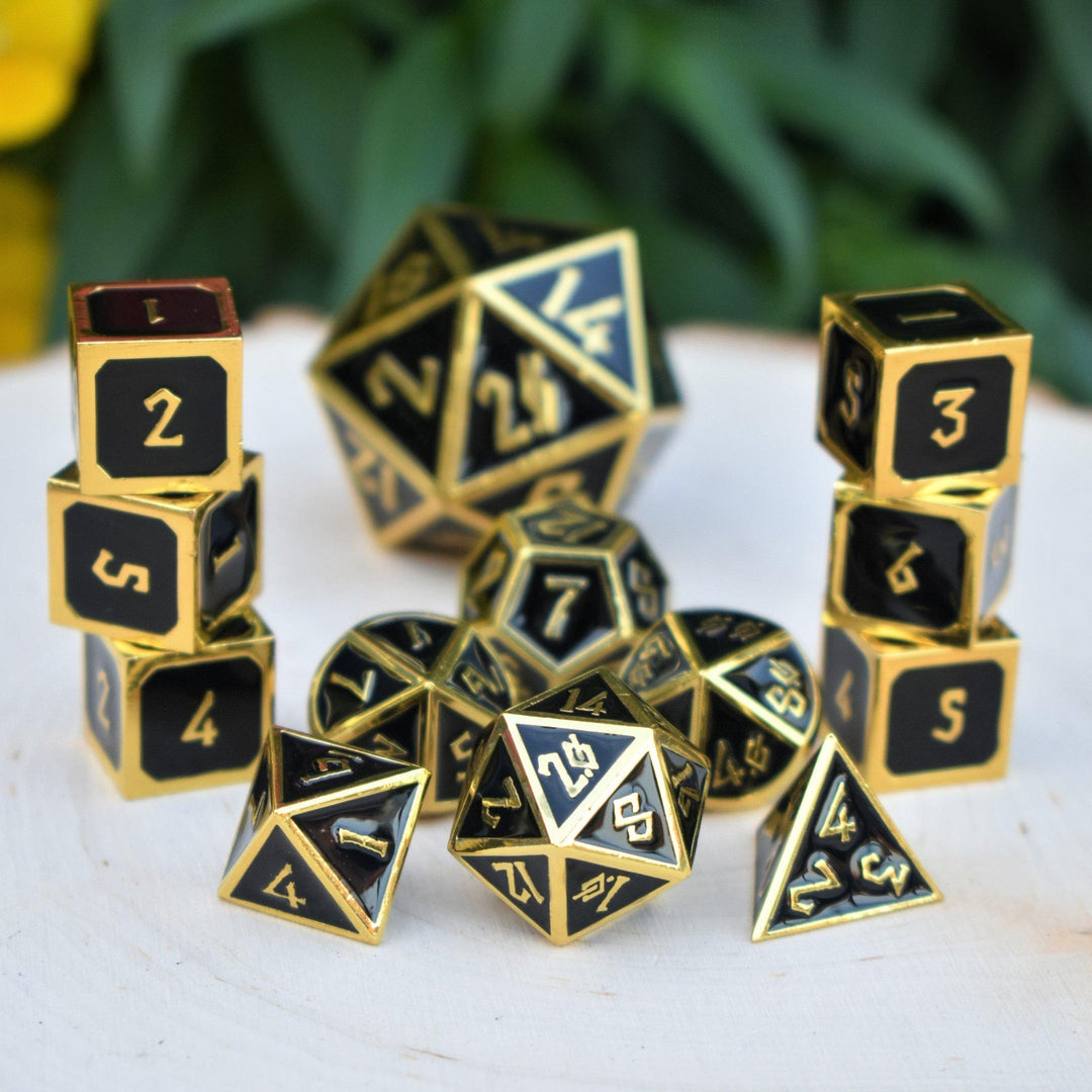A collection of gold and black metal dice with embossed numbers, including various shapes such as a four-sided die, six-sided die, eight-sided die, ten-sided die, twelve-sided die, and twenty-sided die, displayed on a wooden surface with foliage in the background.