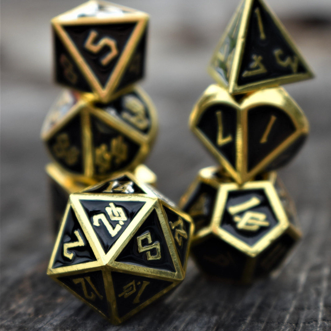 A collection of gold and black metal dice with embossed numbers, including various shapes such as a four-sided die, six-sided die, eight-sided die, ten-sided die, twelve-sided die, and twenty-sided die, displayed on a wooden surface with foliage in the background.