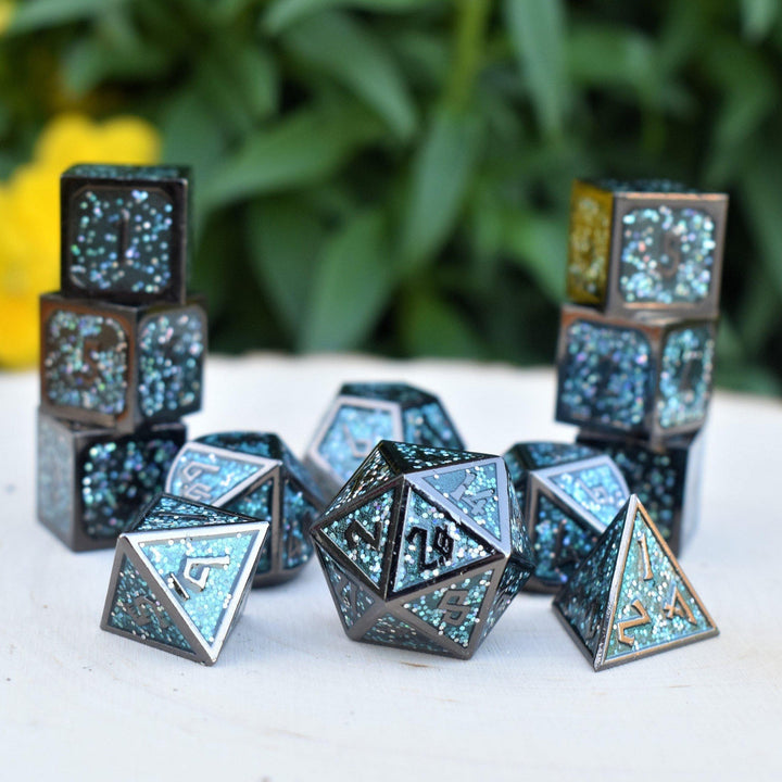 A set of metal dice with embossed numbers, featuring a blue and black color scheme with glittery embellishments, displayed on a white surface with foliage in the background.