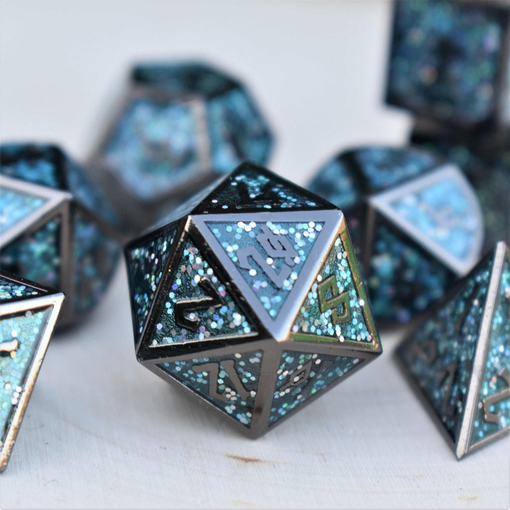 A set of metal dice with embossed numbers, featuring a blue and black color scheme with glittery embellishments, displayed on a white surface with foliage in the background.