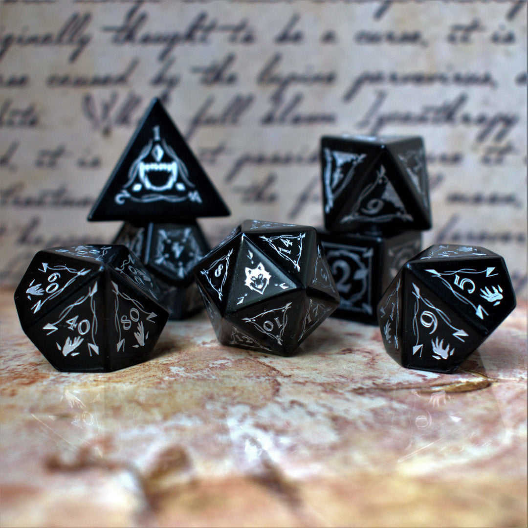 A set of black obsidian stone dnd ttrpg dice with engraved numbers, displayed on a textured surface with a blurred background featuring written text.