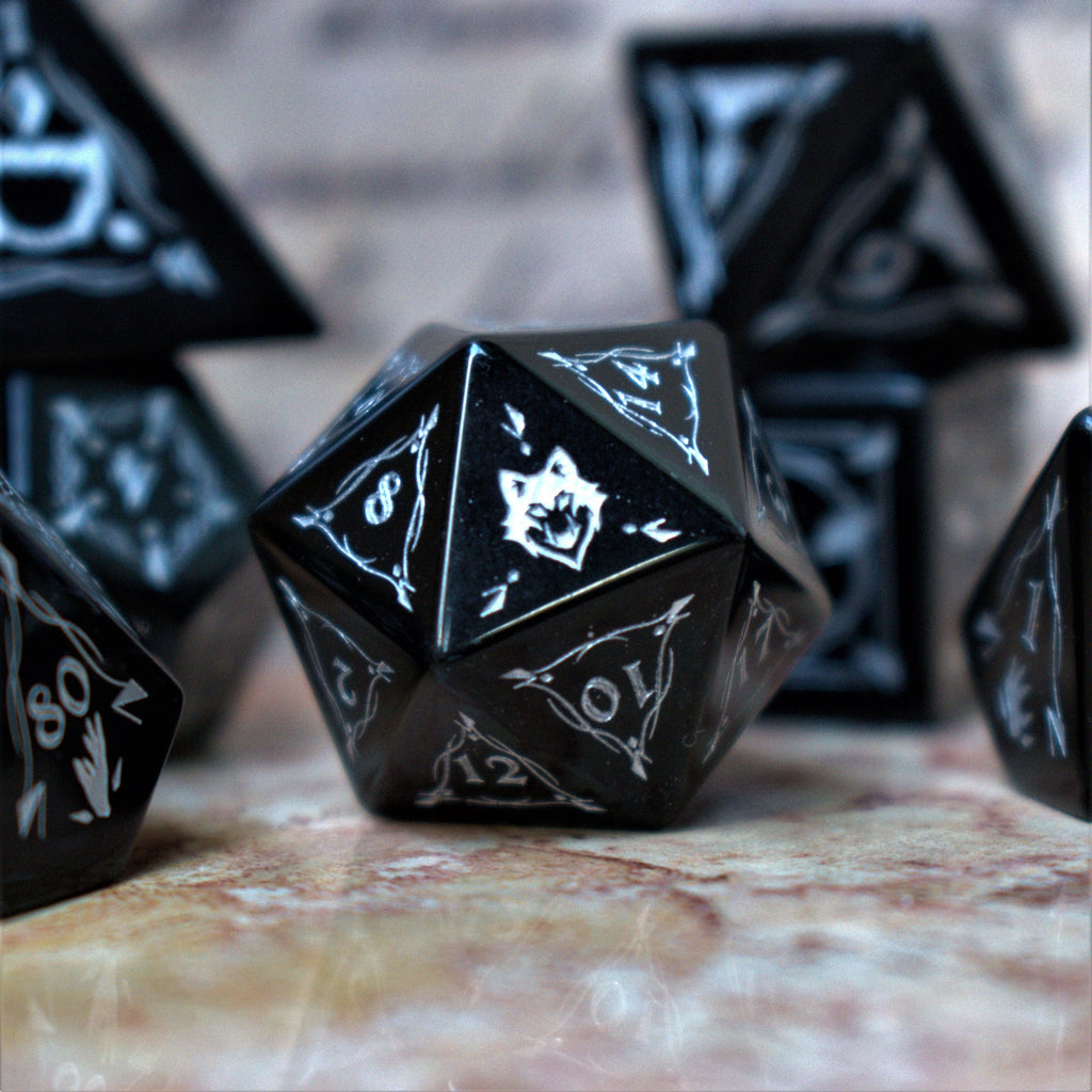 A set of black obsidian stone dnd ttrpg dice with engraved numbers, displayed on a textured surface with a blurred background featuring written text.