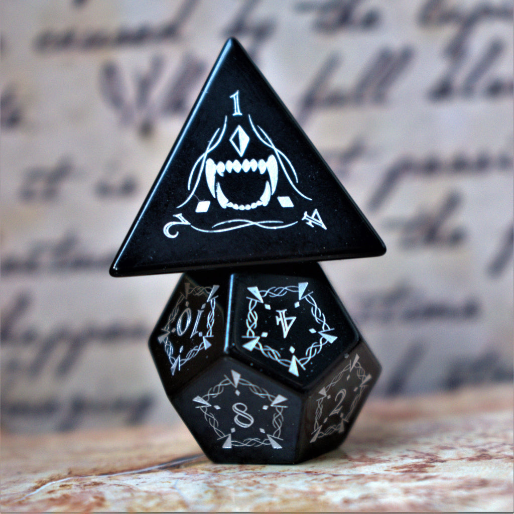 A set of black obsidian stone dnd ttrpg dice with engraved numbers, fangs and fine details, displayed on a textured surface with a blurred background.