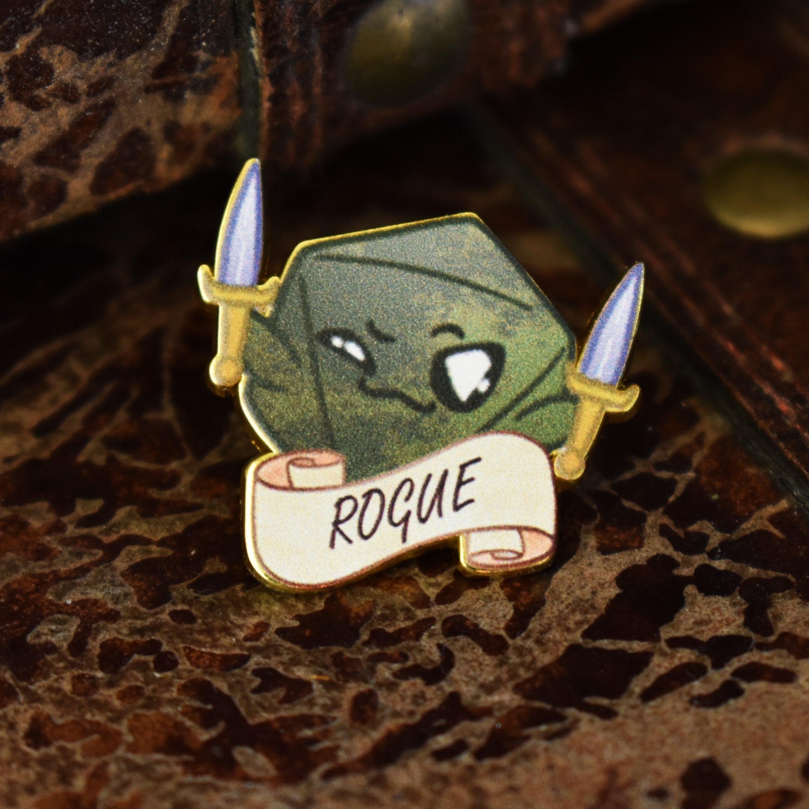 An enamel pin with a character illustration resembling a 'Rogue' class from D&D, featuring a green tone with a sword and the text 'ROGUE' on a banner.