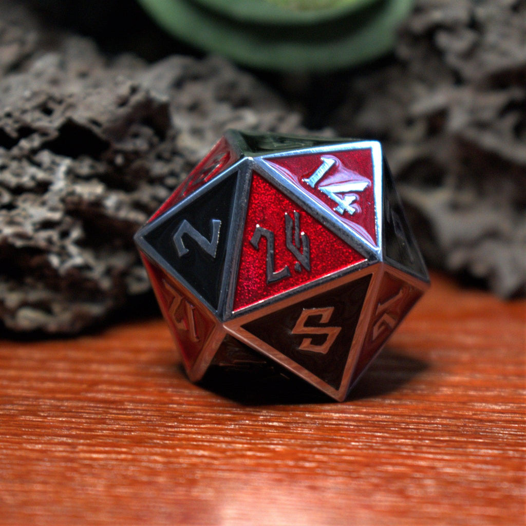A close-up of a scarlet and black metal 35mm D20 dice with silver embossed numbers on a wooden surface.