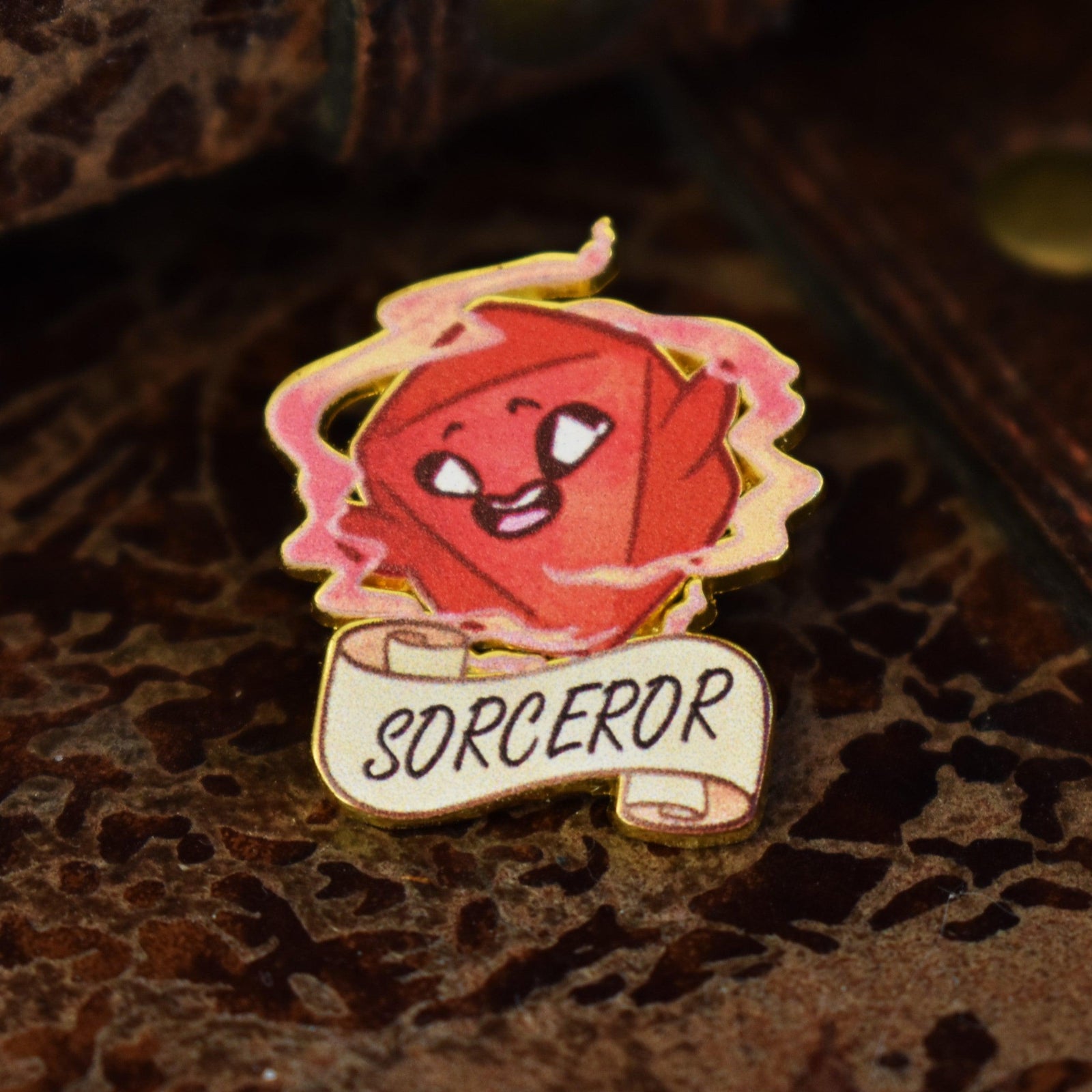 An enamel pin shaped like a sorcerer's dice with the word 'SORCEROR' on it, predominantly red with gold and white accents.
