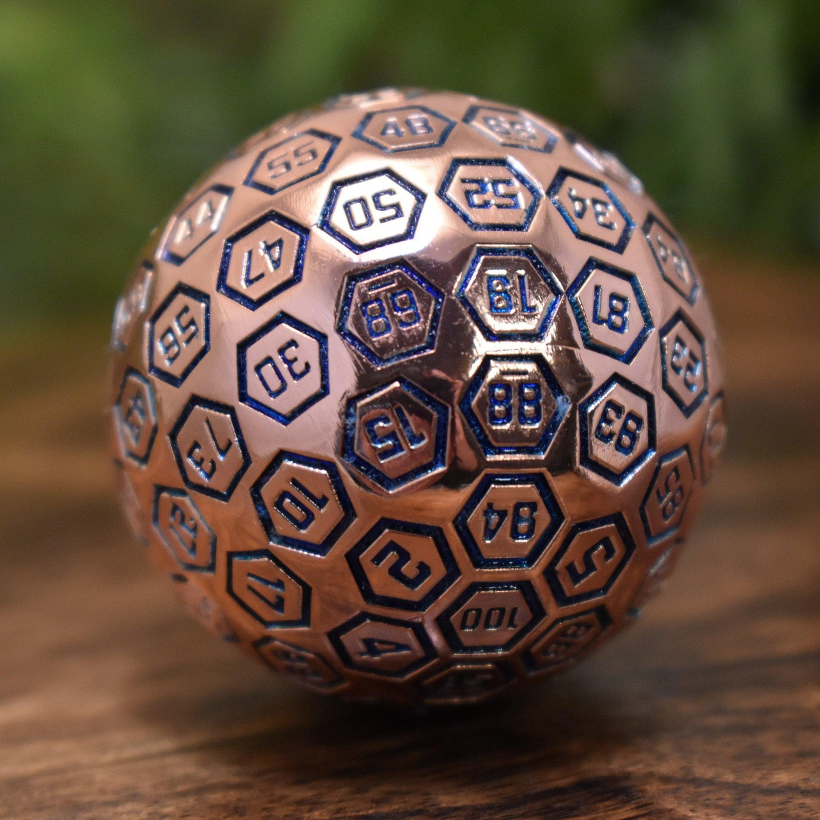 A bronze metal D100 die with blue engraved numbers, displayed on a wooden surface.