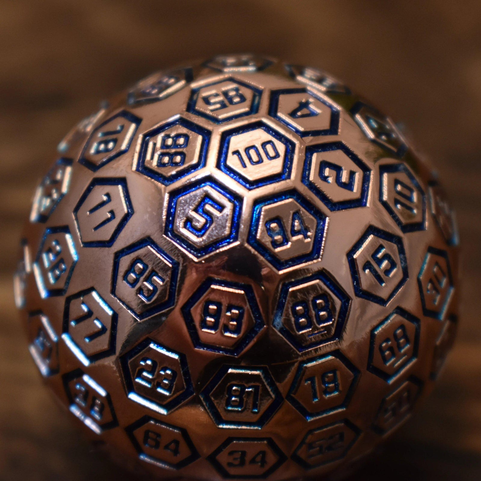 A bronze metal D100 die with blue engraved numbers, displayed on a wooden surface.