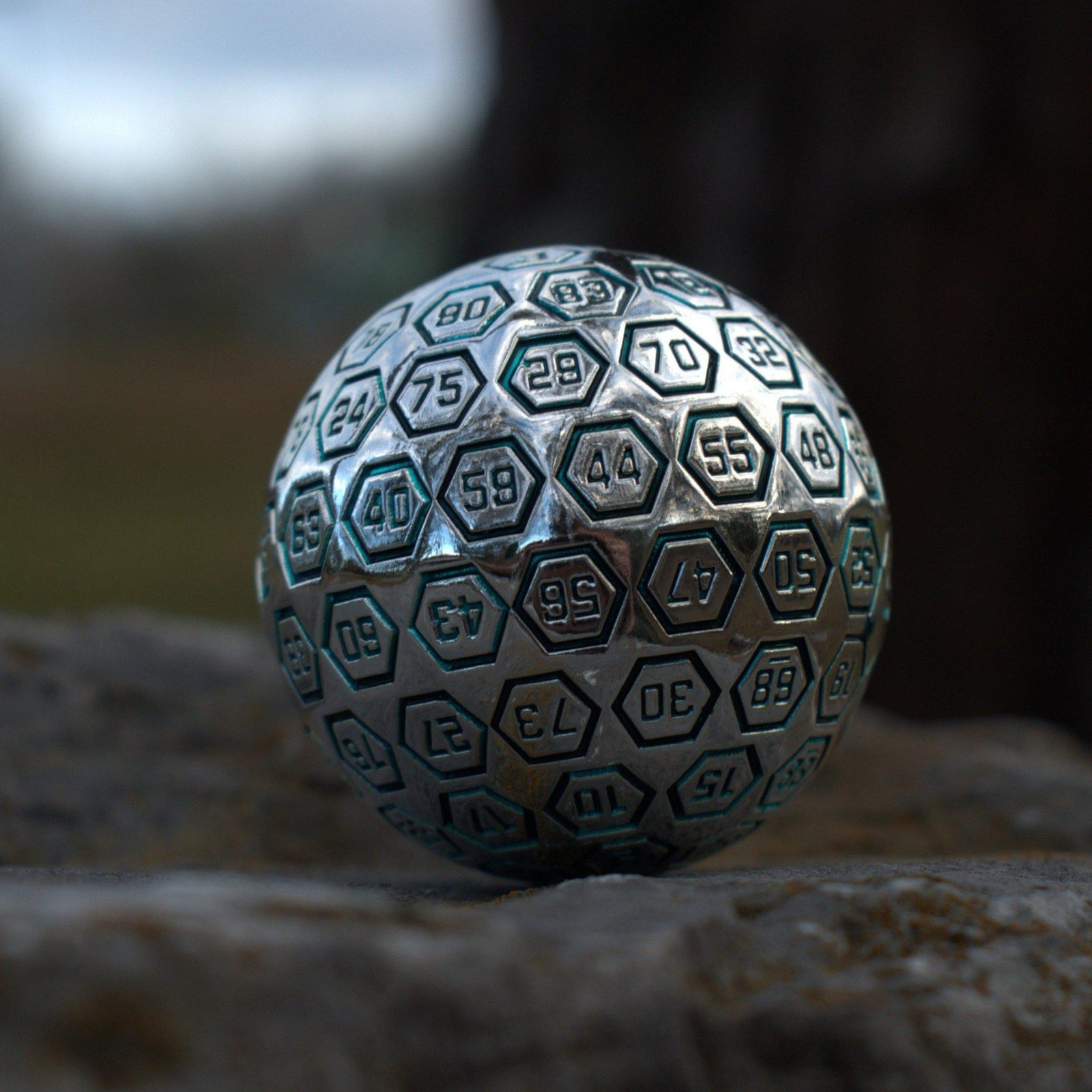 A large silver metal d100 dice with engraved numbers, resting on a rocky surface
