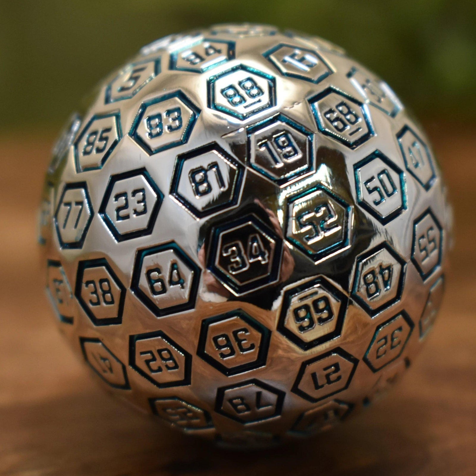 A large silver metal d100 dice with engraved numbers, resting on a rocky surface
