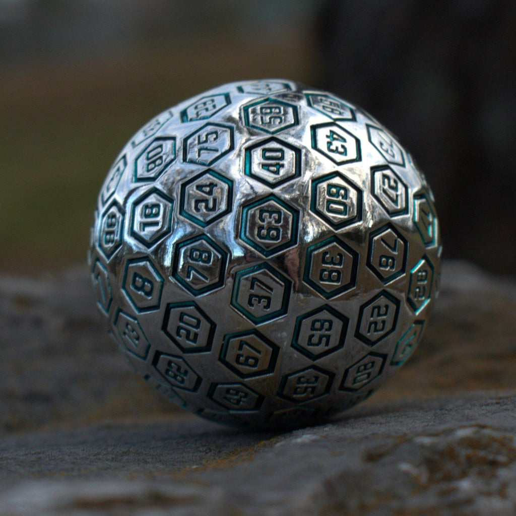 A large silver metal d100 dice with engraved numbers, resting on a rocky surface