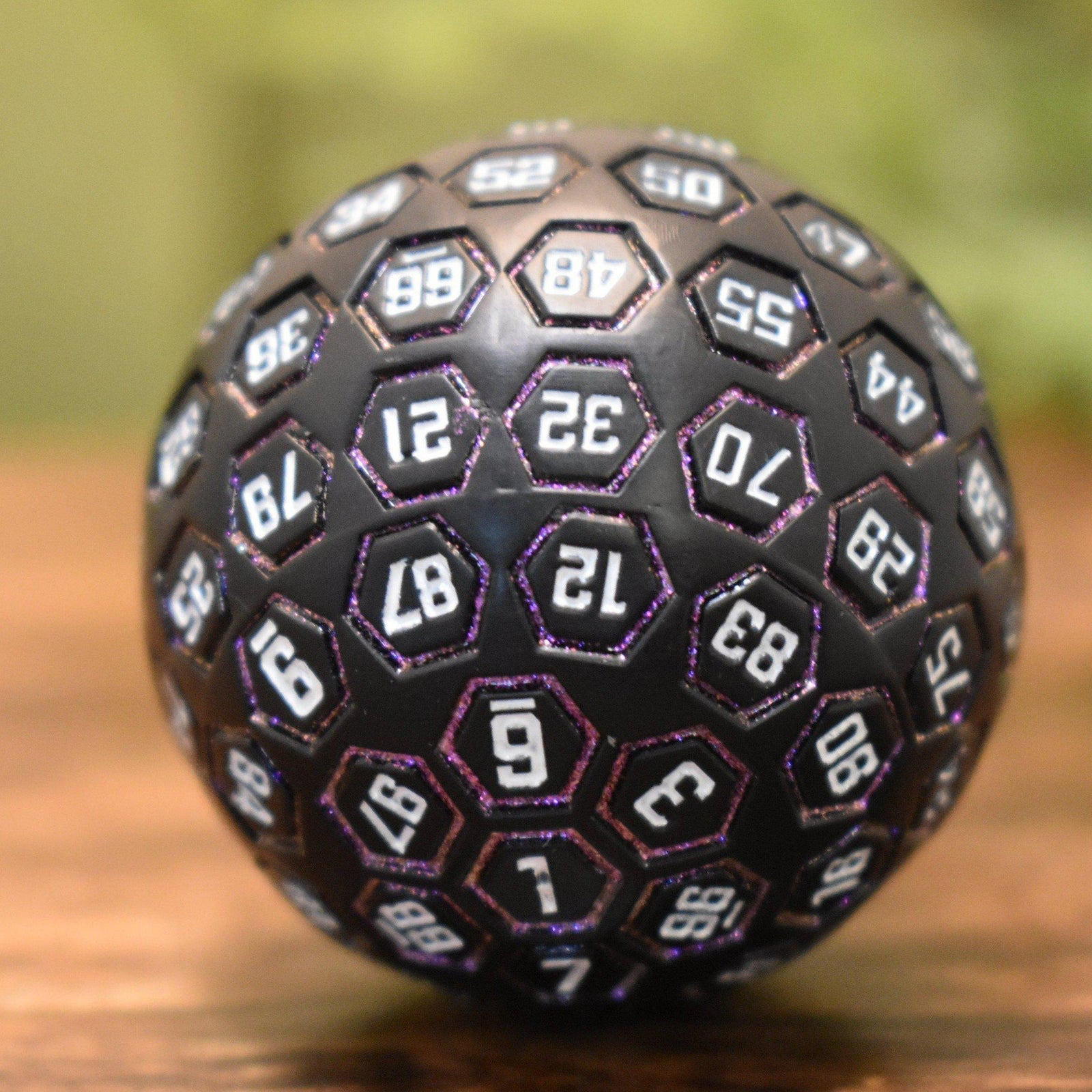 A large, metal D100 dice in purple, white, and black with engraved numbers on each face, displayed on a wooden surface.