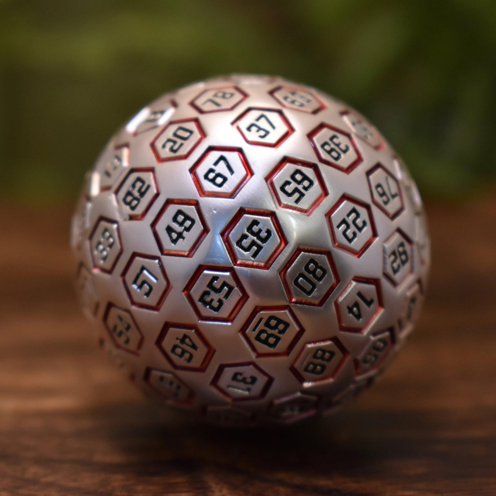 A red and matte silver metal D100 dice with engraved numbers on each face, displayed on a wooden surface.