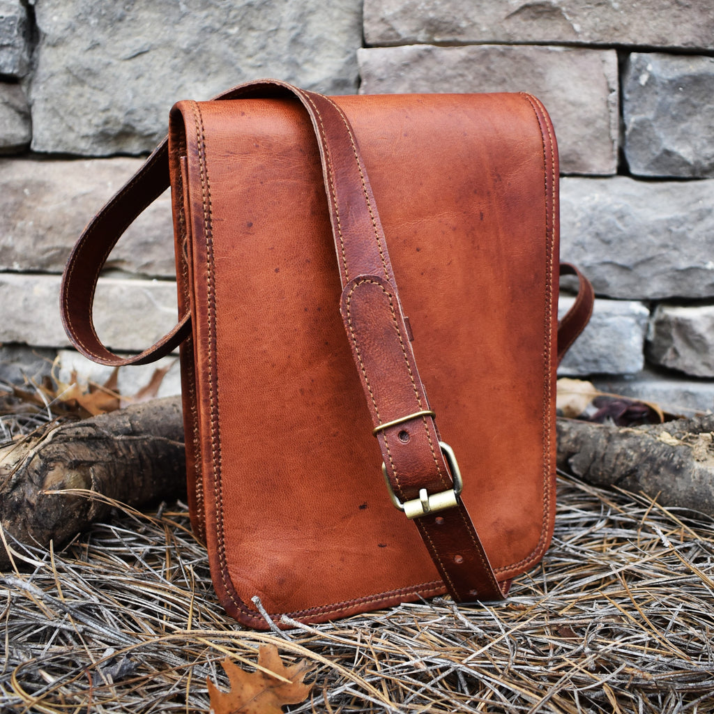 A brown leather satchel with a shoulder strap placed against a stone wall, featuring brass buckles and a flap closure.
