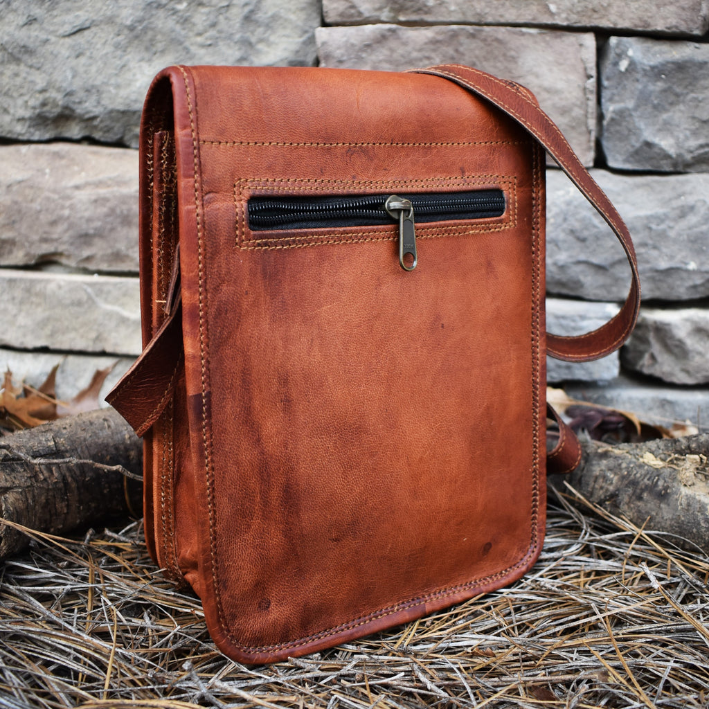 A brown leather satchel with a shoulder strap placed against a stone wall, featuring brass buckles and a flap closure.