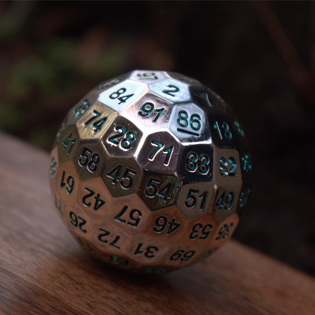 A large silver metal d100 dice with engraved aqua numbers, resting on a wooden surface.