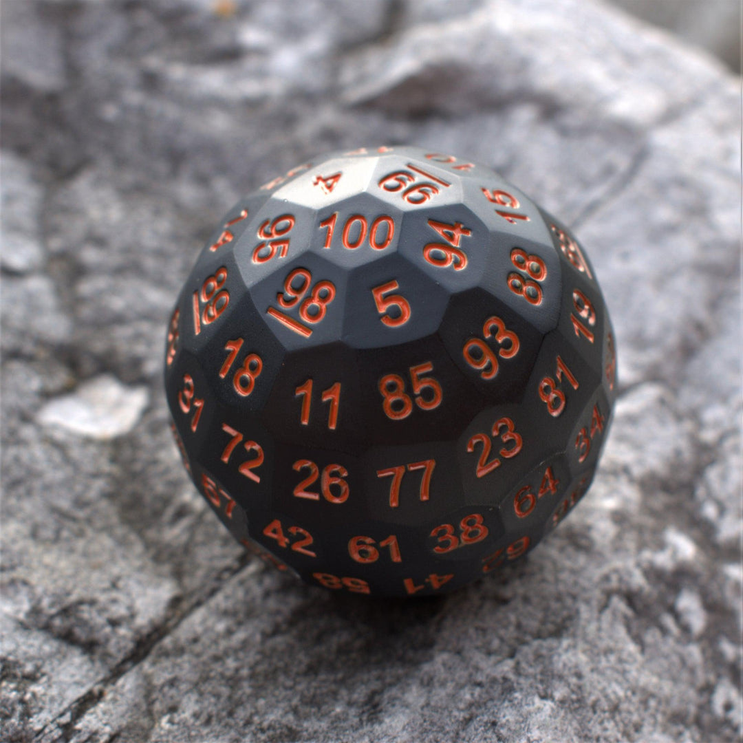 A large black metal dice, a D100, with engraved orange numbers, resting on a rocky surface.