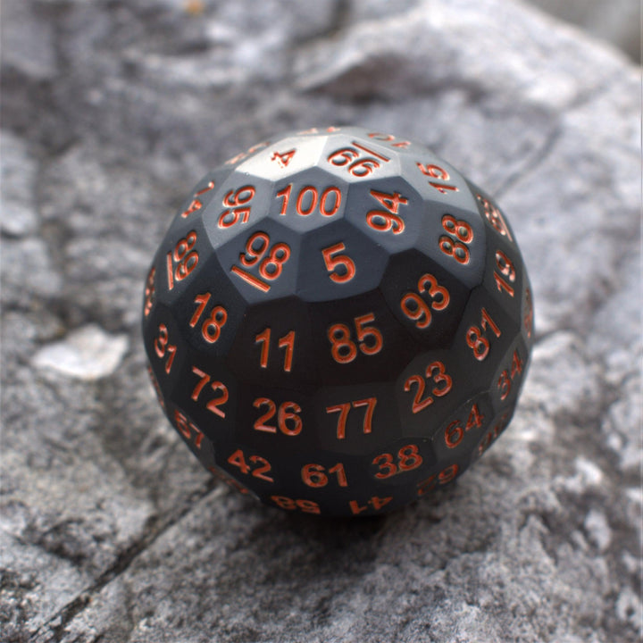 A large black metal dice, a D100, with engraved orange numbers, resting on a rocky surface.