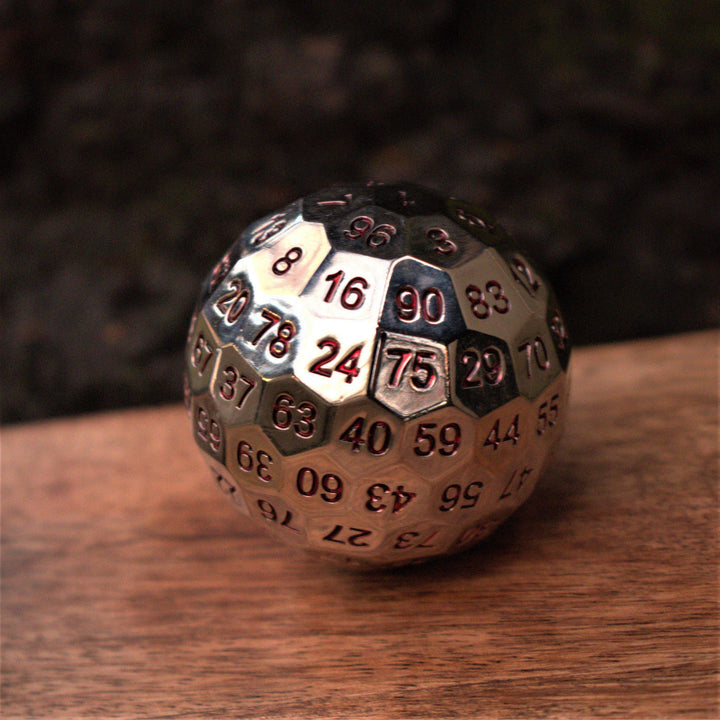 A large silver metal d100 dice with red engraved numbers, resting on a wooden surface for wargaming tabletop gaming board games