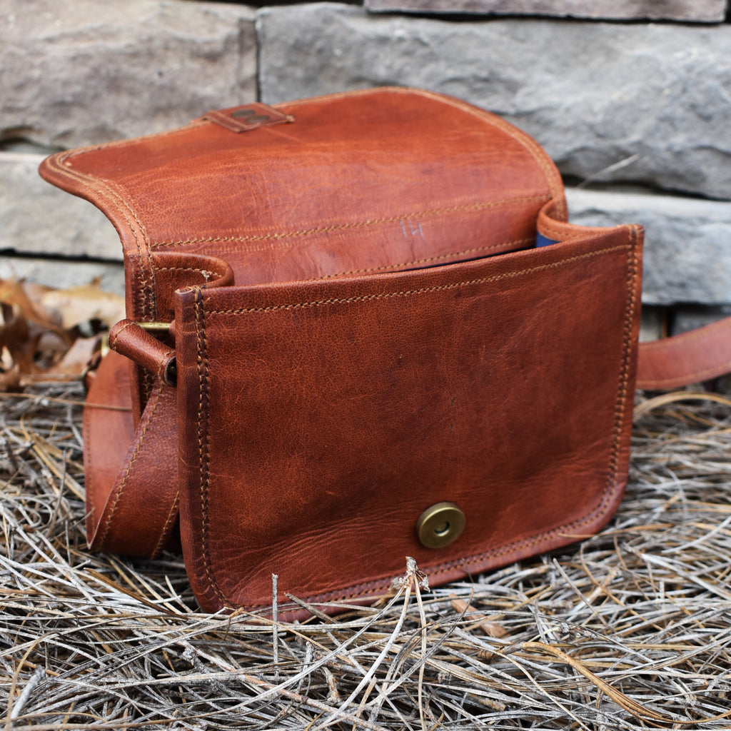A brown leather satchel with brass buckles, an outside zipper, and a shoulder strap, placed against a stone wall with dry leaves around.