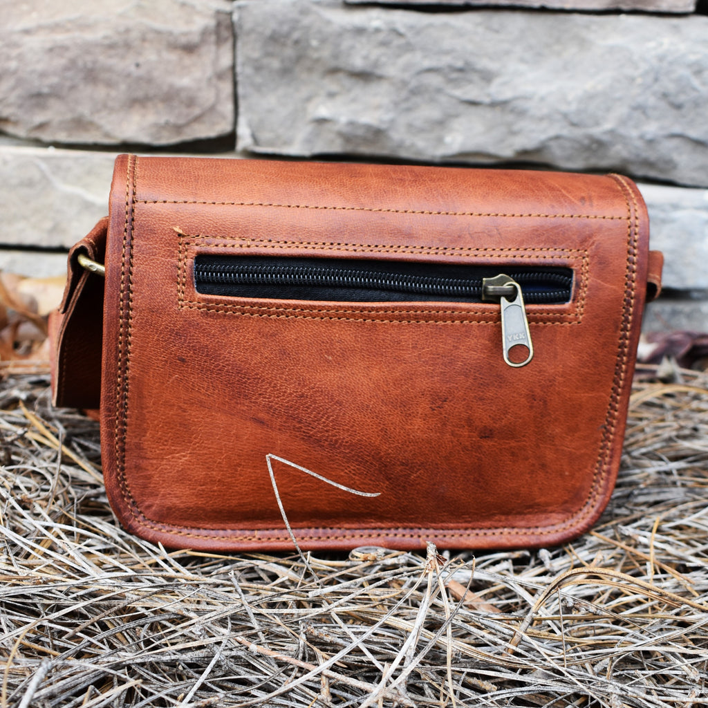 A brown leather satchel with brass buckles, an outside zipper, and a shoulder strap, placed against a stone wall with dry leaves around.