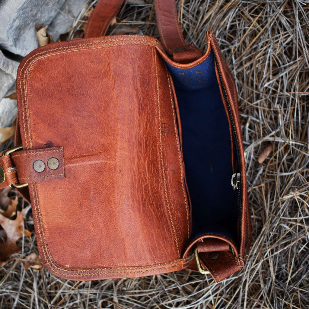 A brown leather satchel with brass buckles, an outside zipper, and a shoulder strap, placed against a stone wall with dry leaves around.