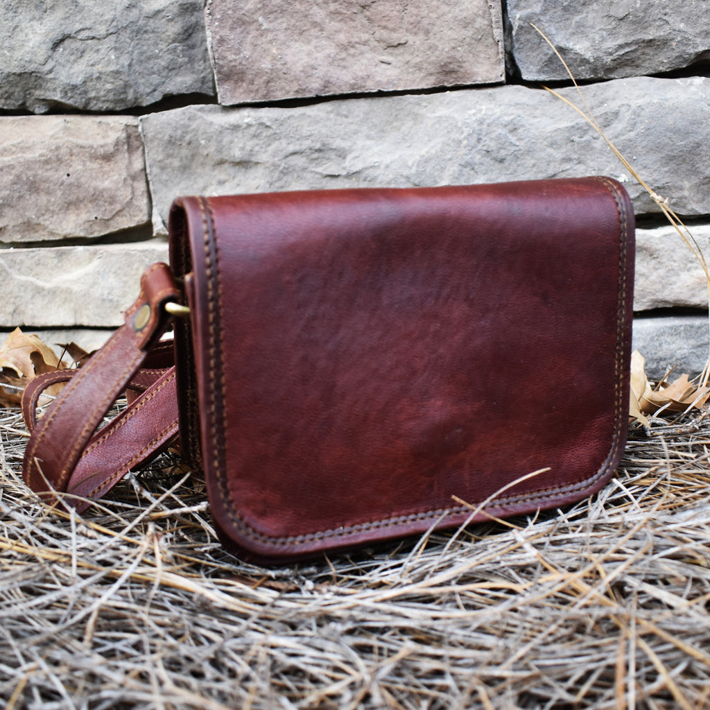A brown leather satchel with a brass buckle placed against a stone wall, with dry grass and leaves around it.