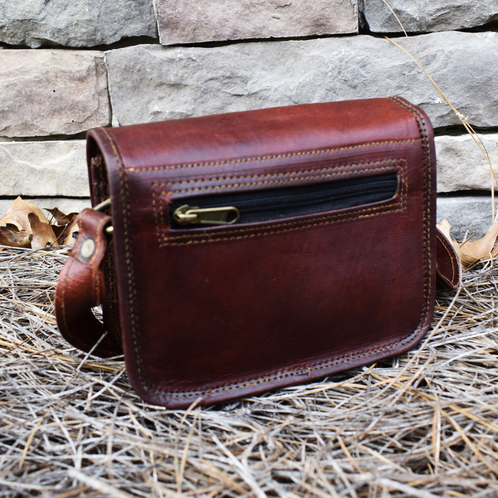 A brown leather satchel with a brass buckle placed against a stone wall, with dry grass and leaves around it.