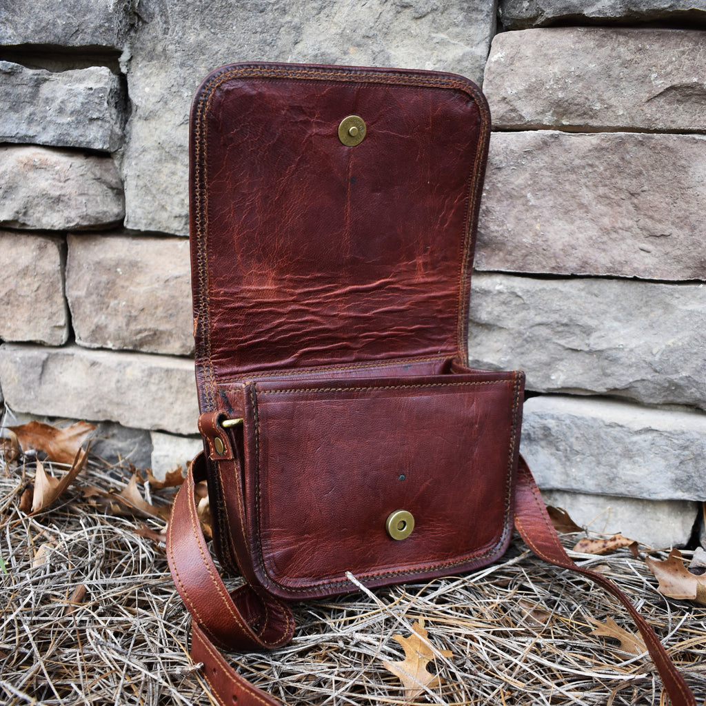 A brown leather satchel with a brass buckle placed against a stone wall, with dry grass and leaves around it.