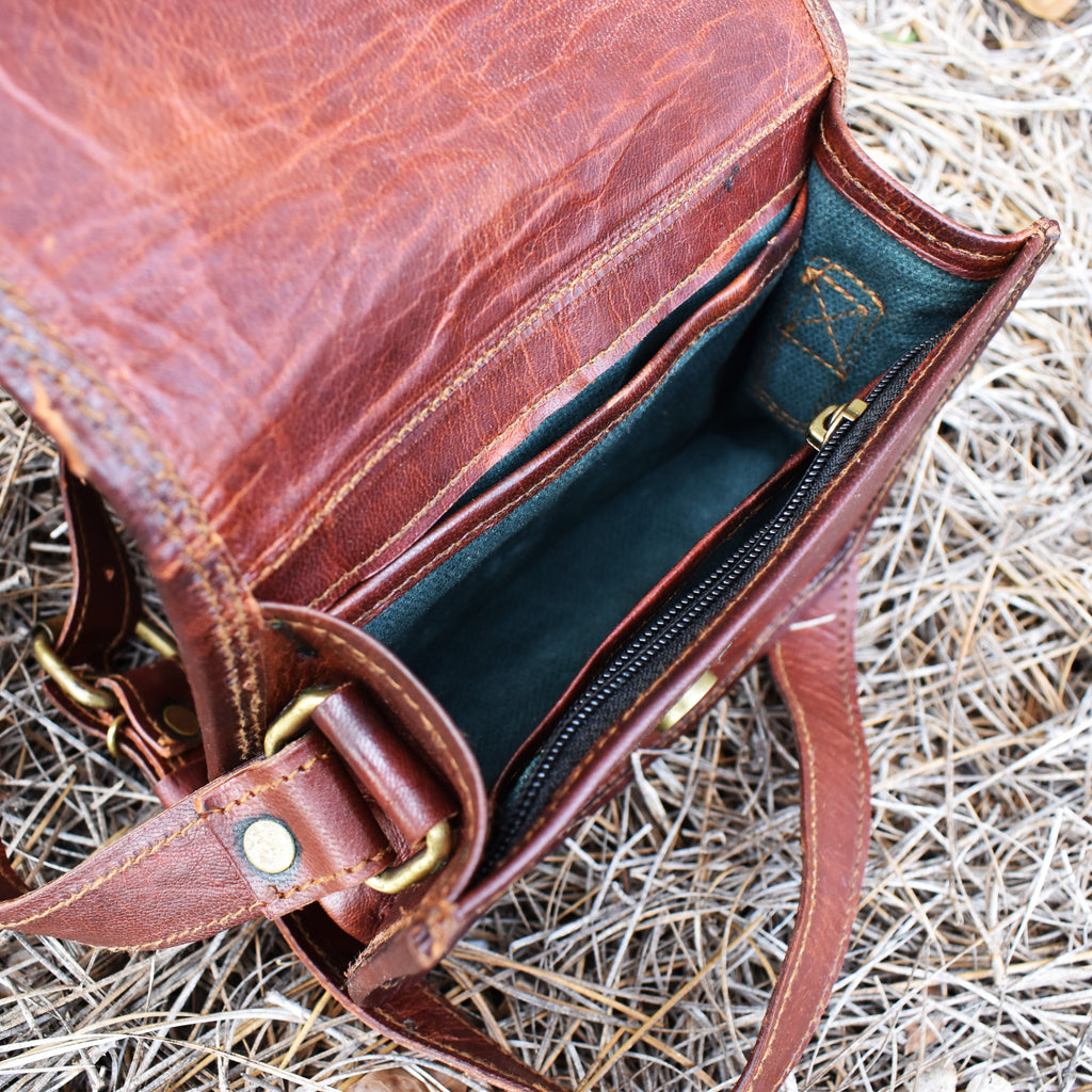 A brown leather satchel with a brass buckle placed against a stone wall, with dry grass and leaves around it.
