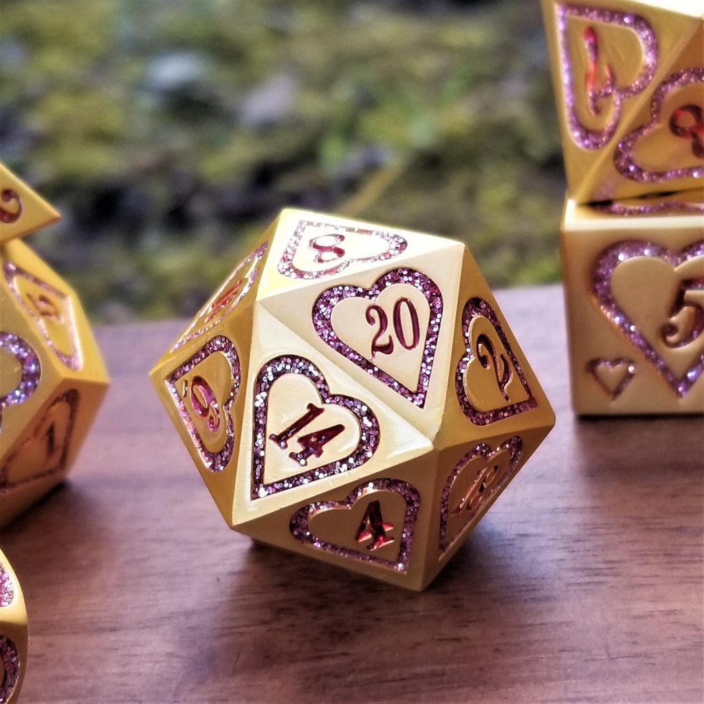 A close-up of a gold metal dice set with pink heart engravings on the faces, placed on a wooden surface with a blurred natural background.